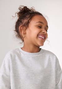 Szürke pulóver kerek nyakkivágással és laza szabással, puha textúrával. Göndör hajú gyermek mosolyog, a háttér egyszínű fehér.