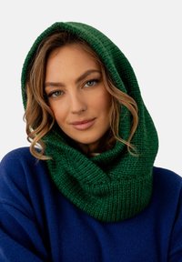 Zielony, dziergany kaptur-kapa z grubego przędzy, charakteryzujący się teksturowanym wzorem i przytulnym, oversizedowym krojem. Noszony z niebieskim swetrem.