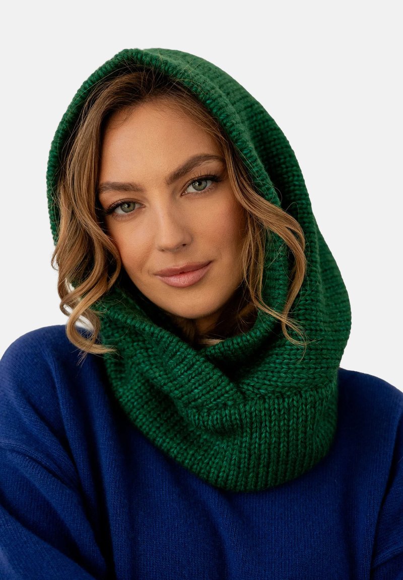 Zielony, dziergany kaptur-kapa z grubego przędzy, charakteryzujący się teksturowanym wzorem i przytulnym, oversizedowym krojem. Noszony z niebieskim swetrem.