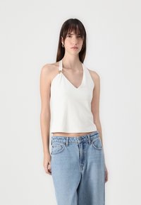Cropped white halter top with a V-neckline, featuring a side knot detail and smooth fabric texture. Paired with loose blue denim jeans.