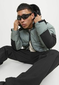 Puffer jacket in light green and black, featuring a zip closure and hood. Black pants visible, adding a textured appearance with large pockets.
