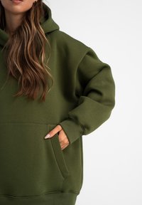 Hoodie vert olive en tissu doux, avec une poche frontale et une coupe décontractée, doté d'un capuchon à cordon de serrage et de poignets côtelés.