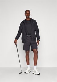 Juodas užtrauktukas su gobtuvu, juoda marškinėliai, juodi šortai su kišenėmis, balti sportbačiai, laikantis golfo lazdą, atrodo moderniai sportiškai.