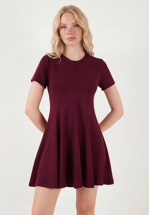 Frau mit langen blonden Haaren, die ein kurzärmeliges, bordeauxfarbenes ausgestelltes Kleid trägt, steht vor einem einfarbigen hellen Hintergrund.