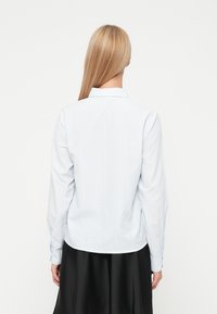 Svetlo-modra, s dolgimi rokavi, srajca s vertikalnimi belimi črtami in šilastim ovratnikom. Gladka tkanina z gumbastimi manšetami. Zadnji pogled.