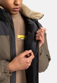 Olijf- en zwarte jas met een fleece-gevoerde kraag, voorzien van een rits en elastische manchetten, die wordt geopend gedragen over een beige shirt.