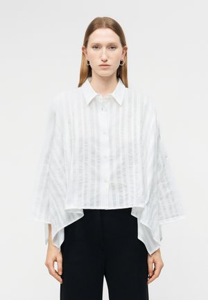 Νεαρή γυναίκα με ίσια ανοιχτό καστανά μαλλιά που φοράει σκουλαρίκια κρίκους και μια άνετη λευκή ριγέ πουκαμίσα με φαρδιά μανίκια και μαύρο παντελόνι.