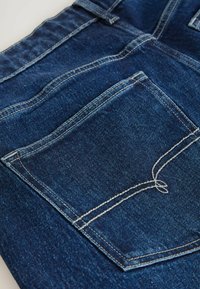 Sötétkék farmernadrág, látható fehér varrással, a hátsó zsebén díszítő dupla varrással, fehér felületen.