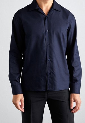 Homme portant une chemise bleu marine foncé à manches longues boutonnée et un pantalon noir, debout devant un fond clair uni.