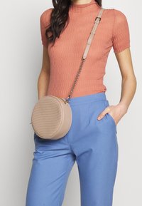 Runde beige Umhängetasche mit gestepptem Muster, Reißverschluss und Kettenriemen, kombiniert mit einem strukturierten Pfirsichoberteil und blauen Hosen.
