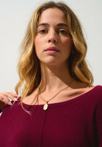 Burgundy ulltröja med en bred halsringning, lager av guldhalsband med en berlock, slät textur och axellångt böljande hår.