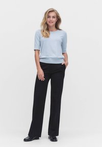 Jasnoniebieski sweter z krótkim rękawem, prążkowany u mankietów i u dołu, połączony z czarnymi spodniami o szerokich nogawkach. Model stoi z rękami w kieszeniach. Czarne buty.