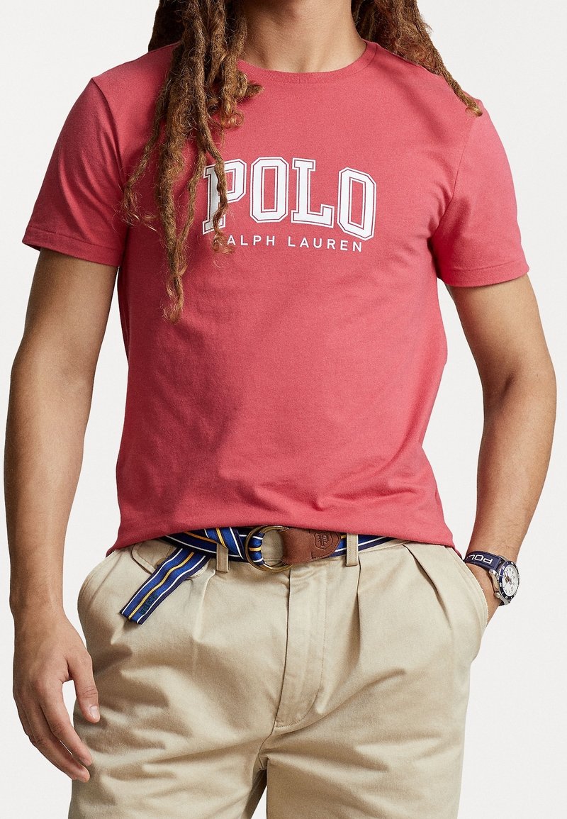 Röd bomullströja med "POLO RALPH LAUREN"-logga i vitt, rund halsringning, korta ärmar, matchad med khakifärgade byxor och ett flerfärgat bälte.
