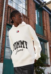 Felpa crema con una grande grafica nera "TOMMY" e un logo più piccolo in rosso e blu. Presenta una tasca frontale e polsini a costine.