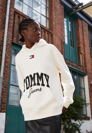 Sudadera crema con un gran gráfico negro de "TOMMY" y un logo más pequeño en rojo y azul. Presenta un bolsillo frontal y puños acanalados.