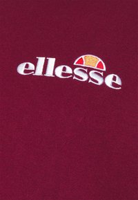 Tessuto di cotone bordeaux con logo ricamato "ellesse" bianco, con un accento a semicerchio rosso e giallo sopra il testo.