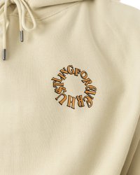Beige hettegenser med en rund oransje grafisk tekst "HUSTLING FOR...". Stoffet har en jevn tekstur, avsluttet med snor og metallikoner.