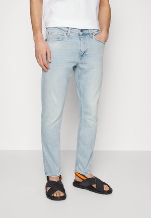 Personne portant un jean bleu clair et des sandales noires avec des brides orange, debout devant un fond clair uni.
