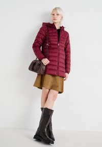 Bordo puffer jakna s kapuljačom, zatvaranjem na zip, i kvadratnim uzorkom; u kombinaciji s smešnom kordura suknjom i smešnim kožnim čizmama do koljena.