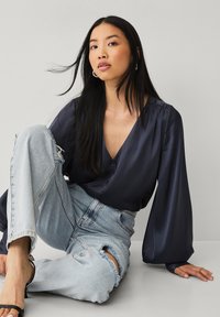 Marineblå satinbluse med lange puffærmer, v-hals og knapdetaljer, parret med lyseblå ripped jeans og sorte sandaler.