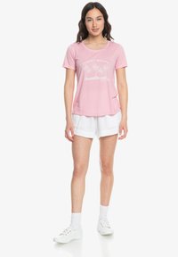 Lichtroze katoen T-shirt met een ronde hals, korte mouwen en een cartoonachtige palmboomafbeelding. Gecombineerd met witte shorts en sportschoenen.
