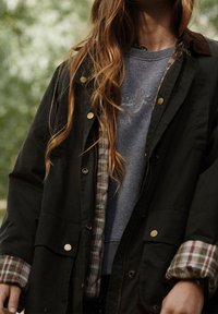 Veste extérieure vert foncé avec des boutons-pression dorés, superposée à un sweat-shirt bleu. Intérieur à carreaux visible au niveau des poignets, tissu texturé.