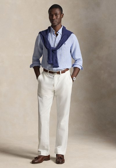 Άνδρας που φοράει μπλε ριγέ πουκάμισο με μπλε σκούρο πουλόβερ περασμένο πάνω από τους ώμους, λευκό παντελόνι, καφέ ζώνη και μοκασίνια, στέκεται με τα χέρια στις τσέπες.