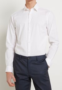 Camisa blanca abotonada con cuello clásico, hecha de tela lisa. Se lleva con pantalones oscuros de cuadros, mostrando un ajuste entallado.