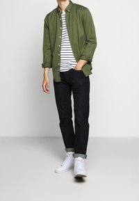 Grön skjorta med knappar över en vit och marinblå randig t-shirt, kombinerat med mörkblå jeans och vita sneakers. Rullade ärmar på skjortan.