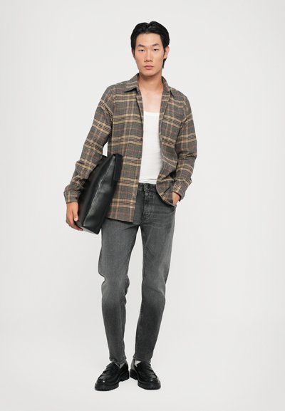 Chemise à carreaux dans des tons terreux, débardeur blanc, jeans gris, chaussures noires, tenant un sac noir. Texture lisse, coupe décontractée et design superposé.