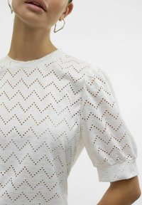 Hvid top med pufærmer, der har et zigzag udskæringsmønster med øjenlids-huller. Glat tekstur, rund hals og detaljerede syninger.