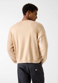 Beżowy sweter z dzianiny o luźnym kroju, opuszczanych ramionach, prążkowanych mankietach i fakturowanym wykończeniu, zestawiony z ciemnymi dżinsami z naszywką z logo.