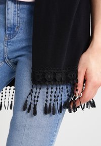 Musta huivi, jossa on rakenteellinen, pitsireunainen helma, jossa koristehelmet. Päällä vaaleansiniset farkut.