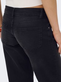 Femme portant un jean en denim noir taille haute avec des poches à l'arrière et un top court en maille blanche, debout devant un fond uni.