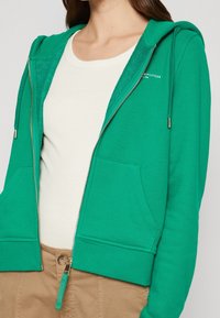 Haut à capuche zippé vert avec une capuche et des poches avant. Le tissu est doux, avec un intérieur texturé. Associé à une chemise crème en dessous.