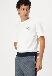 Weißes Baumwoll-T-Shirt mit einem schwarzen Logo-Patch; kombiniert mit grauen gemusterten Jogginghosen, die einen breiten schwarzen Bund und seitliche Streifen haben.