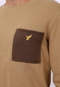 Beige long-sleeve shirt featuring a dark brown chest pocket with an embroidered yellow bird design. Soft, casual fabric texture.