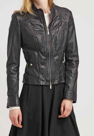 Vrouw draagt een nauwsluitend zwart leren jack met een rits aan de voorkant en gouden drukknopen op de zakken, gecombineerd met een zwarte rok en een witte top met lange mouwen eronder.