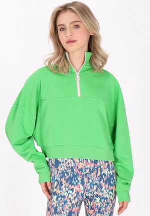 Junge Frau trägt einen leuchtend grünen Sweatshirt mit Reißverschluss und bunte gemusterte Leggings, stehend vor einem weißen Hintergrund.
