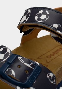 Μαύρη σαγιονάρα με μοτίβο ποδοσφαίρου, ρυθμιζόμενο λουράκι με μπλε πόρπη και καφέ υφασμένη εσωτερική σόλα. Σχεδιασμένη για άνεση και στυλ.