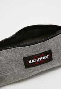 Gray fabric pencil case with a black zipper, featuring an EASTPAK logo patch. Interior is lined in black, showing simple rectangular shape.