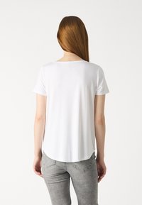 Bela kratka majica s kratkimi rokavi in okroglim izrezom, gladka tekstura in zaobljen rob, v kombinaciji s svetlo sivo prilegajočo se jeans hlačami.