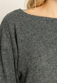 Nahaufnahme einer Person, die einen dunkelgrauen, strukturierten Strickpullover mit einem weiten Ausschnitt trägt, der einen hellen Unterhemdgurt zeigt.
