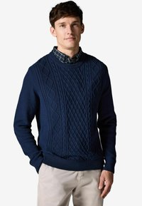 Pulover tricotat bleumarin, cu modele texturate de cablu, guler rotund și manșete și tiv ribbat. Purtați peste o cămașă în carouri.