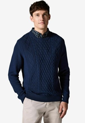 Maglione lavorato a maglia blu navy con motivi a treccia testurizzati, scollo rotondo e polsini e orlo a coste. Indossato sopra una camicia a quadri.
