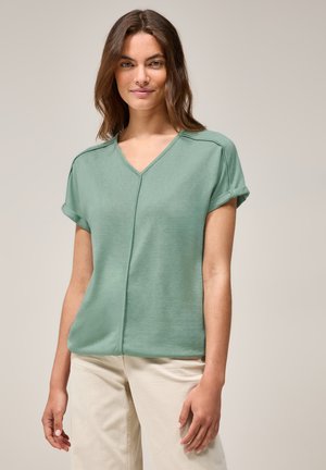T-shirt vert menthe à col en V avec des manches courtes retroussées, présentant un détail de couture sur le devant. Tissu doux et coupe décontractée. Assortie à un pantalon beige clair.