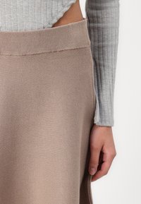 Gonna in maglia beige con vita a costine e una sottile trama. Mano appoggiata all'orlo della gonna. Maglietta a maniche lunghe grigia a coste visibile.