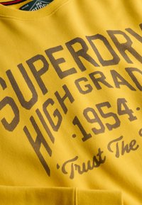 Sweat-shirt jaune avec texte brun foncé "SUPERDRY HIGH GRADE 1954 Trust The" sur le devant.