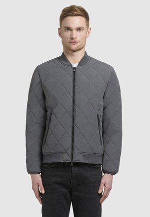 Chaqueta bomber acolchada en gris oscuro. Presenta puños y bajo acanalados, cierre frontal con cremallera y dos bolsillos laterales. Textura suave y acolchada.