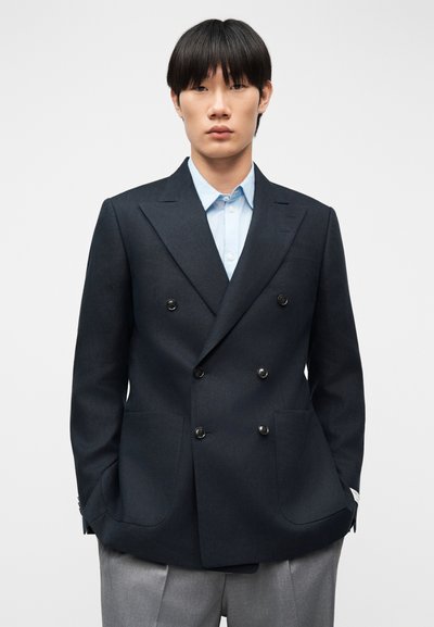 Νέος άνδρας με ίσια μαύρα μαλλιά, ντυμένος με σκούρο σακάκι διπλής φόρμας, ανοιχτό μπλε πουκάμισο και γκρι παντελόνια, κοιτάζοντας μπροστά με τα χέρια στις τσέπες.